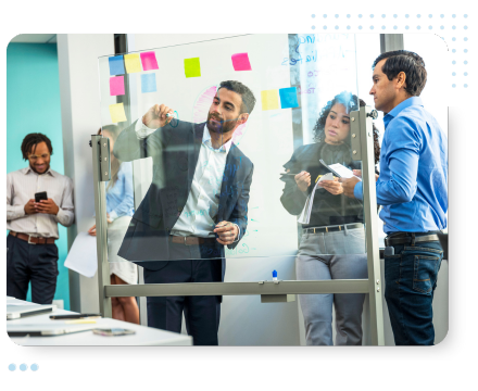 Group collaborates in an office, with a man writing on a glass board. Colleagues watch and take notes. Bright and focused atmosphere.